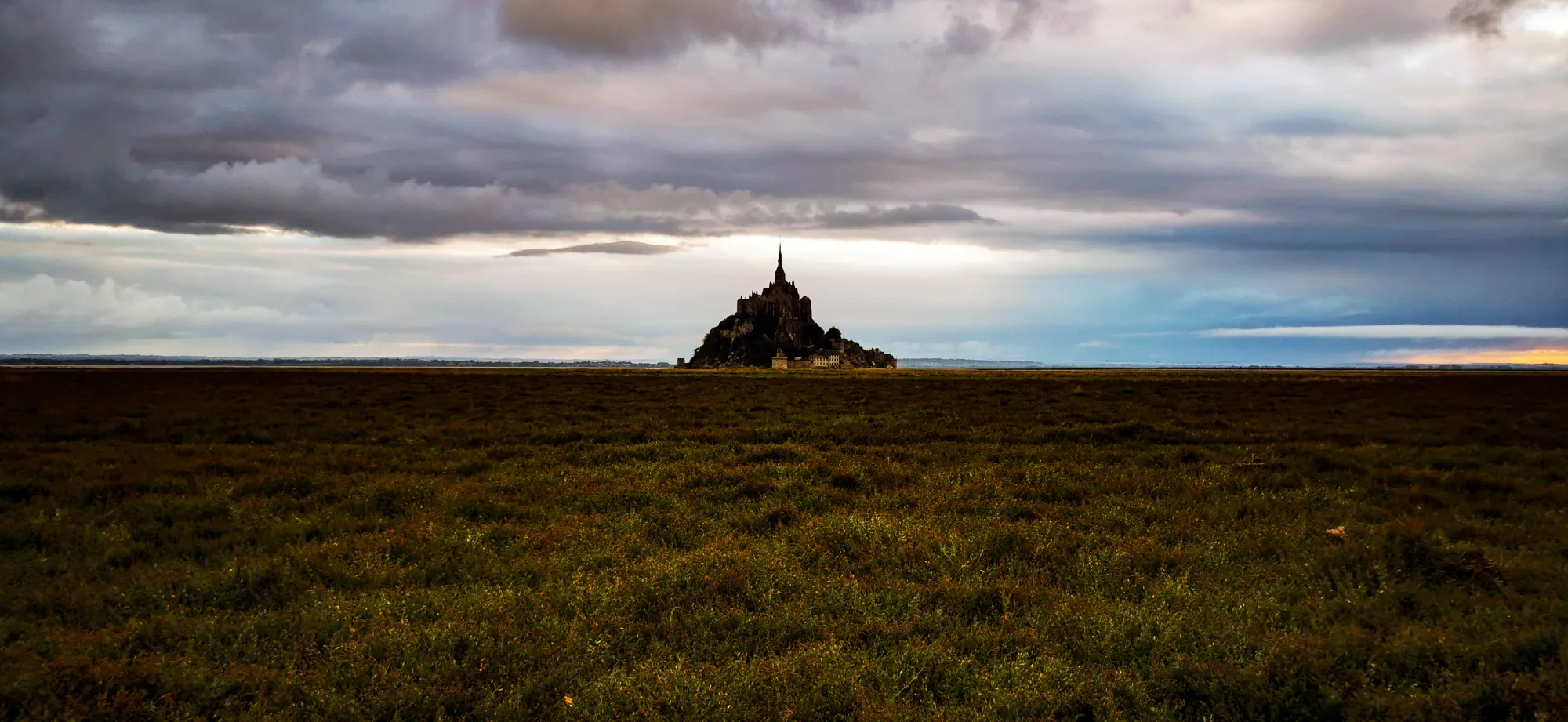 La silhouette du Mont-Saint-Michel au petit matin, sous un ciel de nuages, au bout des herbus